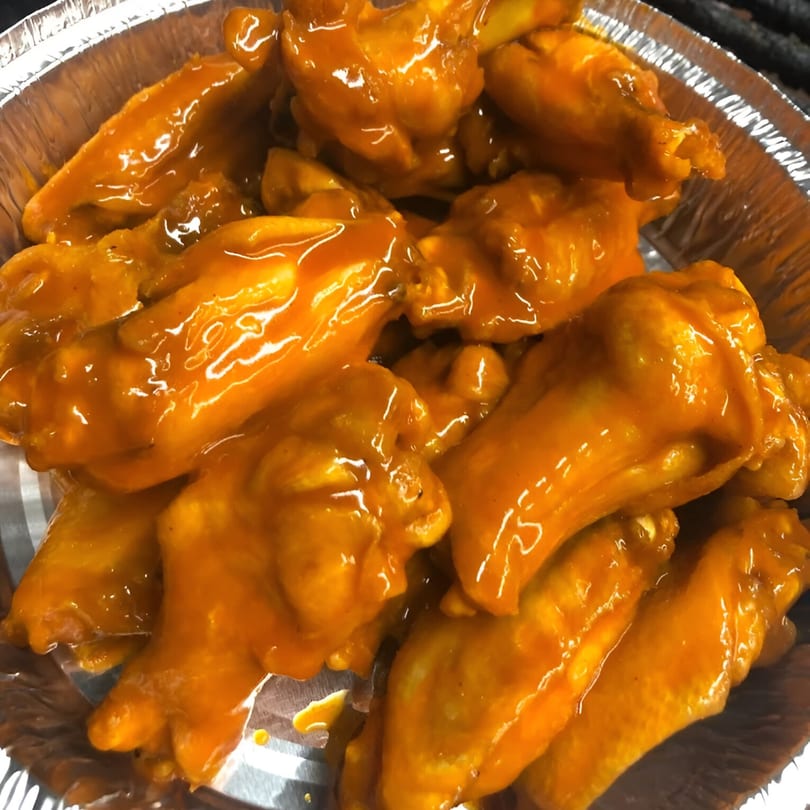 Close-up of FATBOYSBBQ chicken wings, coated in a glistening orange sauce, in a foil container.